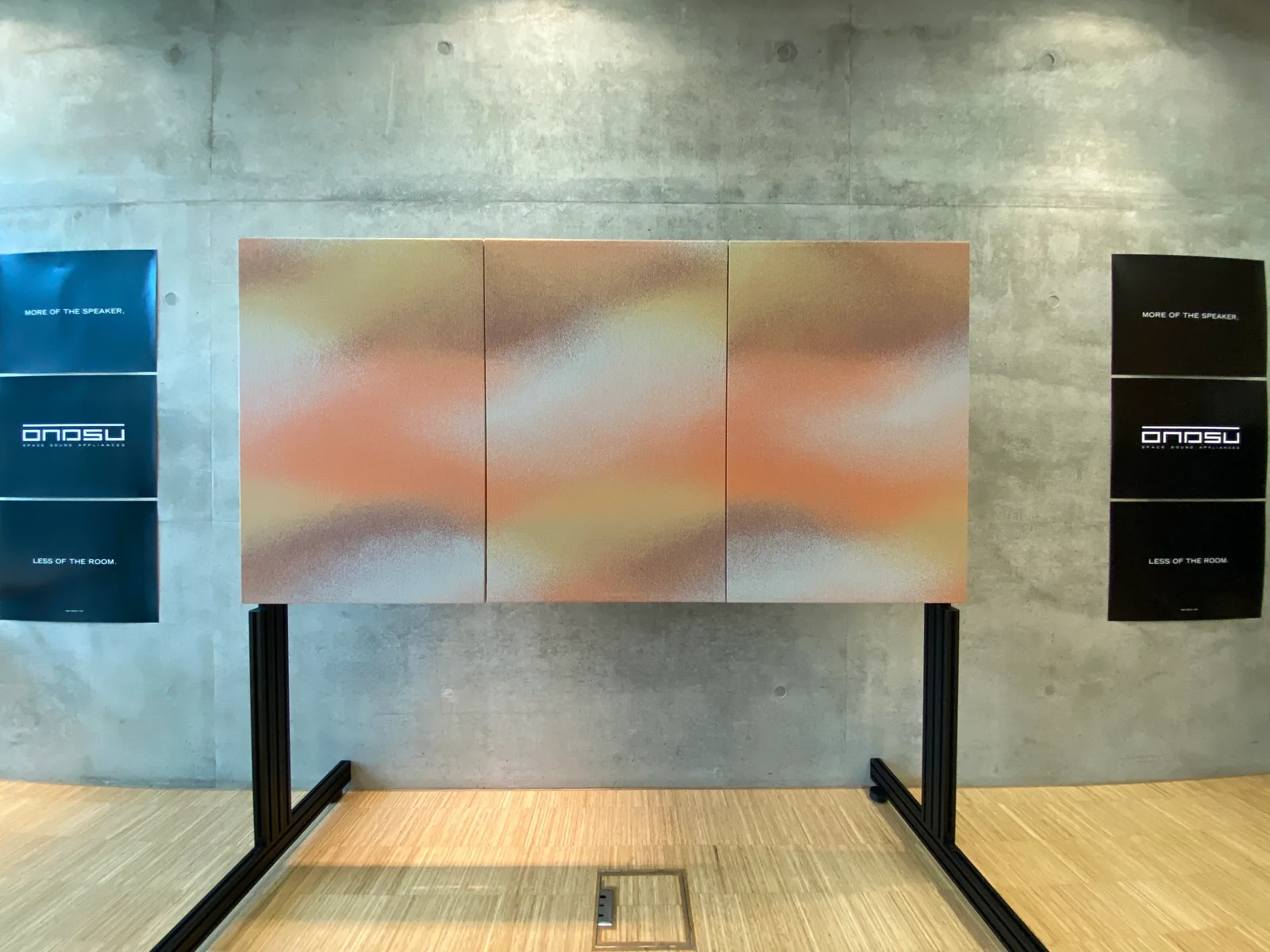 Three acoustic panels with a continuous orange, cream and pale blue textile pattern, on a black freestanding support between ONDSU posters.